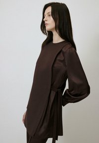 Tunică lungă cu mâneci lungi din satin maro, cu un croi lejer, detalii cu nod pe lateral și un luciu subtil. Decolteu rotund și siluetă fluidă.