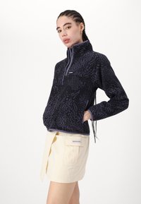 Fleece-pullover i svart med lila abstrakt mönster, halvzip fram, hög krage och sidofickor, kombinerad med en ljusfärgad kjol.