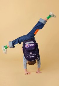 Dark purple backpack with star pattern, front pocket, and floral badge; denim pants with orange and yellow accents; green socks and white shoes.