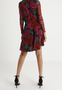 Blumenkleid mit schwarzem Grund und roten sowie pinken Blumen, langen transparenten Ärmeln und weitem Rock. Kombiniert mit schwarzen High Heels.