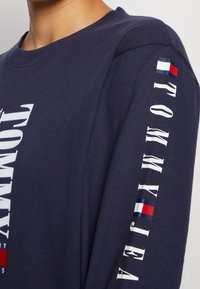 Námořnické tričko s dlouhým rukávem vyrobené z bavlny, s kulatým výstřihem, s výrazným bílým textem a tříbarevným logem na pravém rukávu.