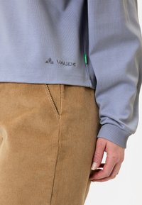 Hellblaues Langarmoberteil mit gerippten Bündchen und dezentem Vaude-Logo. Kombiniert mit beigen Cordhosen, die vertikale Rippung aufweisen.