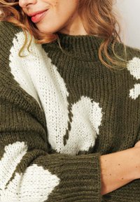 Pull en tricot avec une base en vert olive texturé, orné de motifs abstraits blancs, col montant et manches larges.