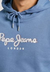 Modrá mikina s kapucí s logem "Pepe Jeans London" a stahovacími šňůrkami svázanými do mašle na krku, nošena osobou.