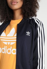 Námorně modrá mikina na zip s bílými pruhy na rukávech, s malým bílým logem Adidas. Kombinováno s jasně oranžovým tričkem s velkým logem.