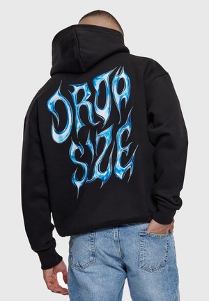 Person iført sort hættetrøje med blå stiliseret "DROP SYNC" tekst på ryggen og lyseblå jeans, hånd i lommen, vendt væk.