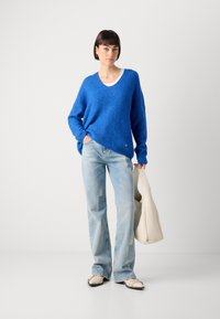 Bright blue knit sweater with a V-neck, paired with light blue flared jeans. Model holds a white bag with a smooth texture and rounded shape.