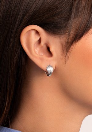 Gros plan sur une oreille droite portant une boucle d'oreille en forme de coquillage argenté, avec des cheveux brun foncé repliés derrière l'oreille.