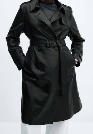 Trench-coat ceinturé en cuir synthétique noir, à double boutonnage, larges revers et poches, porté sur une chemise foncée et un pantalon blanc.