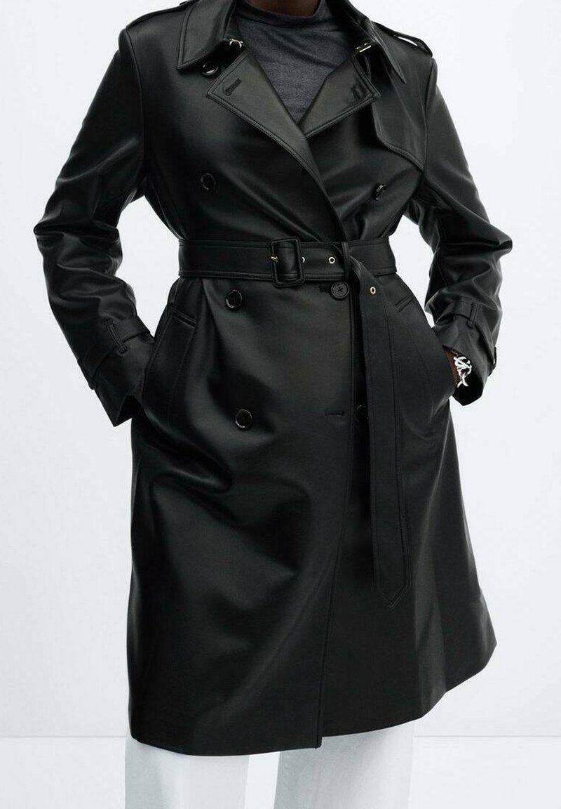Trench-coat ceinturé en cuir synthétique noir, à double boutonnage, larges revers et poches, porté sur une chemise foncée et un pantalon blanc.