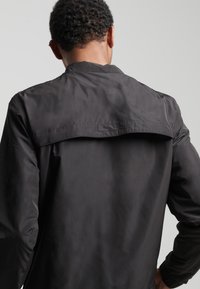 Chaqueta ligera negra con una textura suave, que presenta un detalle de solapa en la parte posterior. El diseño incluye mangas largas y un cuello.