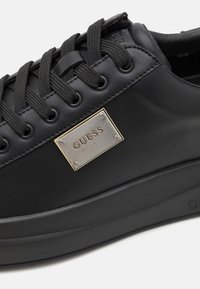 Zapatilla negra con parte superior de cuero liso, cordones texturizados y una placa de metal con el logo grabado. Cuenta con una suela de goma gruesa.