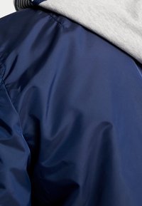 Mörkblå jacka med en glänsande yta, har en grå inre foder och visar en slät textur samt sömdetaljer vid kragen.