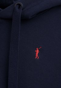 Felpa navy con tessuto testurizzato e una piccola figura rossa ricamata che tiene una mazza da golf sul petto. Regolazione del cordino sul cappuccio.
