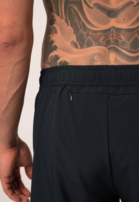 Černé sportovní šortky s elastickým pasem, které mají malou zipovou kapsu na boku a hladkou texturu. Tělesné umění viditelné na zádech.