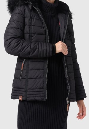 Personne portant une veste matelassée noire avec une capuche doublée de fourrure et un pull à col roulé côtelé noir, les mains ajustent la fermeture éclair de la veste.