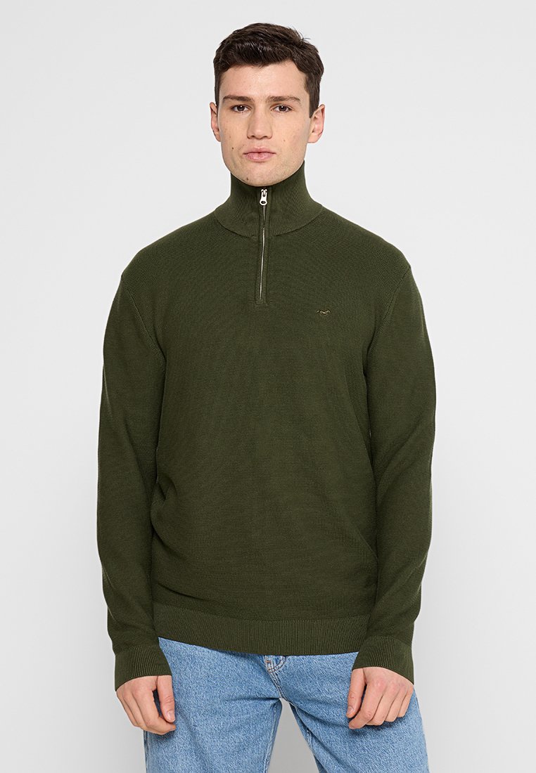 Dunkelgrüner Reißverschluss-Pullover mit hohem Kragen, gerippter Textur, langen Ärmeln und kleinem Logo auf der Brust. Kombiniert mit hellblauen Jeans.