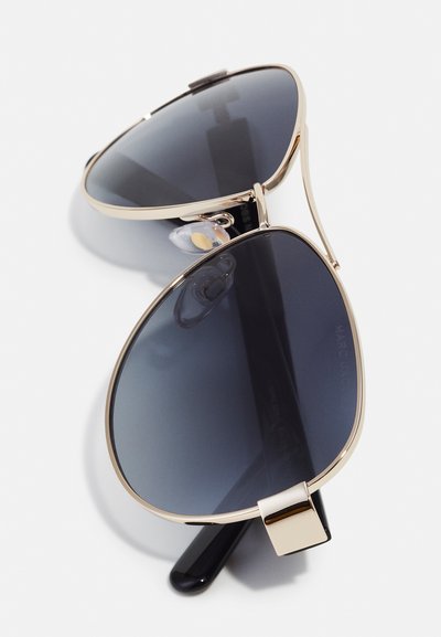 Metallische Flieger-Sonnenbrille mit dunkel getönten Gläsern, goldenen Akzenten und klaren Nasenpads, die eine elegante, überdimensionale Tropfenform aufweist.