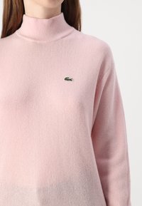 Rosa tröja med hög krage och ribbad textur. Har en liten grön broderad krokodil-logga på bröstet.