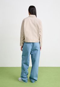 Beige cotton jacket with a collar, paired with loose blue trousers. Simple design and texture, standing on a green surface.