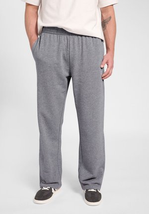 Man draagt losse grijze joggingbroek met zijzakken en zwarte sneakers, staat met handen in de zakken tegen een effen achtergrond.