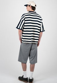 Homme portant une chemise à manches courtes blanche et bleu marine rayée, un short gris à fines rayures arrivant aux genoux, des chaussettes blanches, des chaussures noires et une casquette beige, vu de dos.