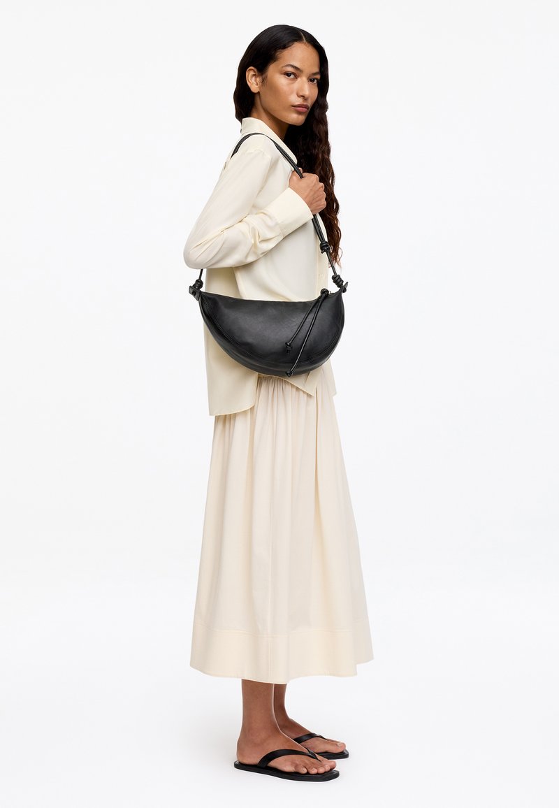 Femme portant un chemisier et une jupe crème, des sandales noires, et tenant un grand sac à bandoulière noir en forme de croissant sur son épaule droite.