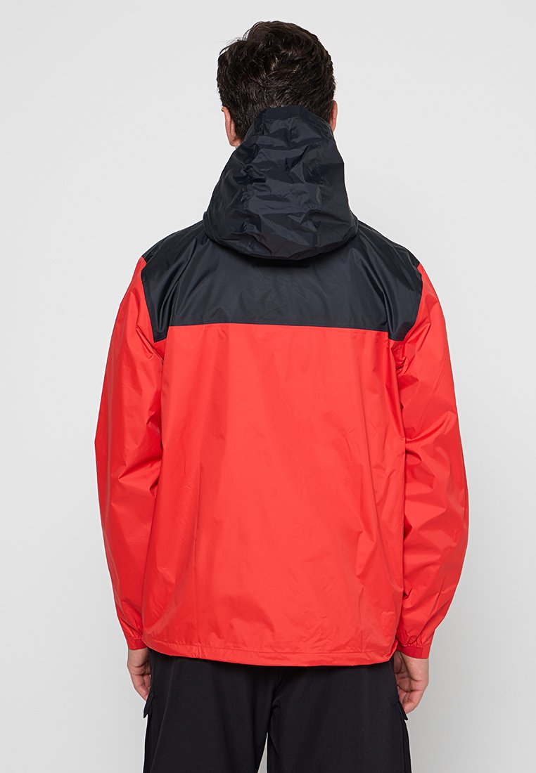 Uomo che indossa un impermeabile con cappuccio rosso e nero, di spalle, con pantaloni neri contro uno sfondo chiaro e semplice.