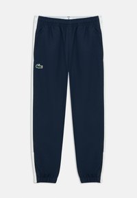 Tmavomodré športové nohavice s bielymi bočnými pruhmi, elastickým pásom a malým zeleným logom Lacoste na ľavom stehne.