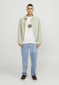 Jachetă bej deschis peste un hanorac gri cu logo, purtată cu blugi deschis la culoare și adidași negri. Croială slim, design minimalist.