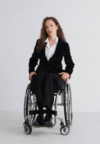 Blazer de terciopelo negro sobre una camisa blanca, combinado con pantalones negros y sentado en una silla de ruedas plateada, mostrando un diseño elegante y características funcionales.