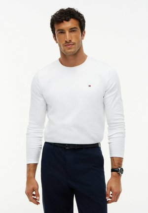 Homme portant une chemise blanche à manches longues avec un petit logo sur la poitrine, un pantalon foncé et une montre-bracelet, debout devant un fond blanc.