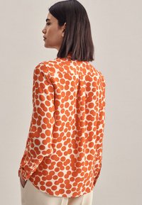 Blusa a maniche lunghe in crema chiaro con grandi pois arancioni. Presenta un colletto, polsini con bottoni e un dettaglio a pieghe sul retro.