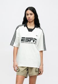 Bela majica z kratkimi rokavi z črnim ovratnikom in tremi črnimi črtami na rokavih. Spredaj je velik napis "ESPN" in logotip.