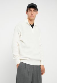 Bela pletena zip-up jopica z rebrasto teksturo, v kombinaciji s sivimi nabranimi hlačami. Model nosi črno kapo, roke v žepih.