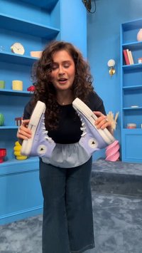 Ženska z kodrastimi lasmi prikazuje par vijoličnih visokih superg Converse v modri sobi s policami in dekorativnimi predmeti.