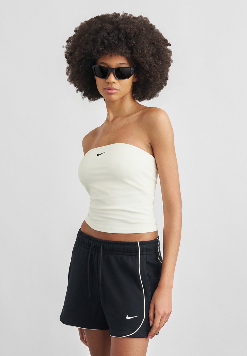 Frau mit großen lockigen Haaren, die eine schwarze Sonnenbrille, ein weißes trägerloses Nike-Oberteil und schwarze Nike-Shorts vor einem schlichten Hintergrund trägt.