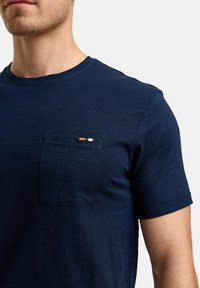 Tricou bleumarin cu mânecă scurtă, realizat dintr-un material texturat, având un buzunar pe pieptul stâng cu un accent subțire în trei culori: portocalie, albă și neagră.