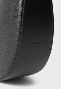 Melna ādas rokassoma ar akmeņainu tekstūru, izliekto formu un iegravētu logotipu. Gludie akcenti un redzamā šuvju detaļa uzlabo dizainu.