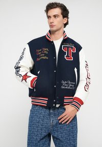 Chaqueta varsity azul marino con mangas de cuero blanco, texto bordado, acentos de estrella y cuello y puños de canalé rojo y blanco a rayas.