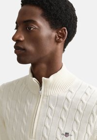 Biely sveter na zips s pleteným vzorom, rebrovaným golierom a logom na hrudi. Textúra je mäkká a hustá.