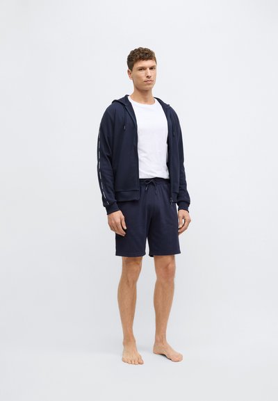 Νavy μπλέ φούτερ με φερμουάρ και κορδόνι, σε συνδυασμό με ναυτικά σορτς. Και τα δύο κατασκευασμένα από μαλακό ύφασμα. Το μοντέλο στέκεται ξυπόλητο μπροστά σε απλό φόντο.