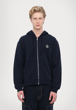 Marineblå zip-hoodie med frontlommer, snoretræk i hætten og broderet logo på brystet. Fremstillet af et blødt bomuldsblandingsstof.