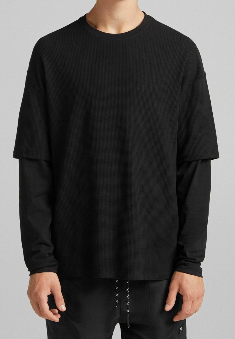 Osoba v několika vrstvách, se černým dlouhým rukávem pod krátkým rukávem texturovaného černého trička a s černými kalhotami s vzorovanými šňůrkami.