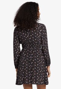 Žena s kudrnatými vlasy, oblečená v dlouhých černých šatech s malými růžovými, modrými a žlutými květinovými vzory, viděná zezadu.