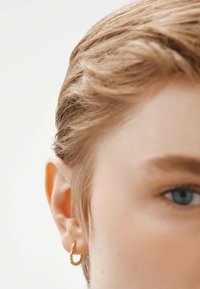 Pendiente de aro dorado sujeto a una oreja de piel clara. Cabello rubio corto y texturizado y un ojo azul son parcialmente visibles, contra un fondo claro.