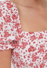 Weißes Oberteil aus Stoff mit rotem Blumenmuster, pufferten Ärmeln, quadratischem Ausschnitt und geraffter Textur. Verfügt über Rüschensäume und ein smocktes Design.