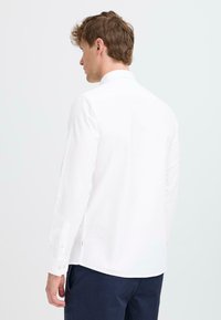 Camisa blanca de manga larga hecha de algodón, con cuello clásico, puños con botones y dobladillo recto, vista desde la parte posterior.
