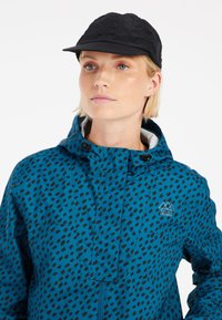 Mėlynas vandeniui atsparus švarkas su juodu abstrakčiu raštu, turintis aukštą apykaklę, užtrauktuką priekyje ir logotipą. Pora su juodu kepurėle.