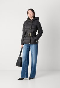 Zwarte pufferjas met capuchon, gequilt ontwerp, gouden hardware accenten, tailleband. Gecombineerd met blauwe broek met wijde pijpen en een zwarte tote bag.